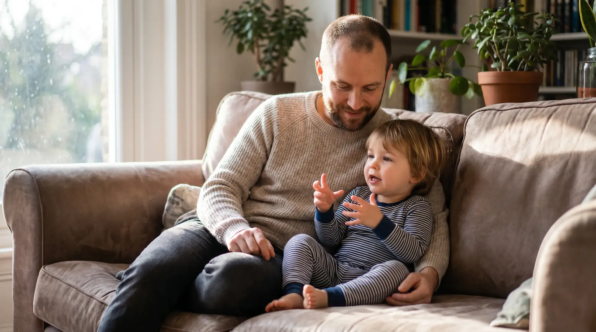 An adult sitting on a couch with a child on their lap. The adult is smiling and looking at the child, who is gesturing with their hands. Potted plants and a bookshelf are visible in the background, and sunlight streams through a window, creating a warm and cozy atmosphere.