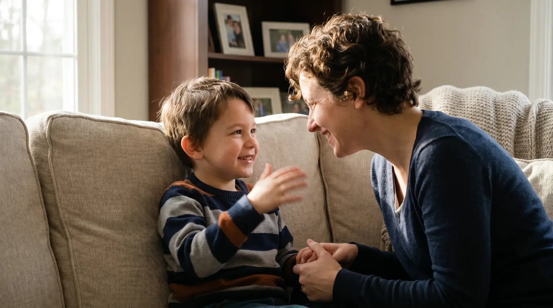 A woman and a young boy sitting on a sofa, smiling and interacting. The boy is making a playful gesture with his hand. Framed photos and a window are visible in the background, creating a cozy, family-like setting.